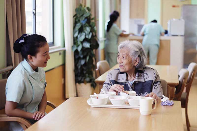 An elderly woman enjoys a meal while engaging in a warm conversation with a caregiver at a senior care facility, reflecting care and companionship in a nurturing environment